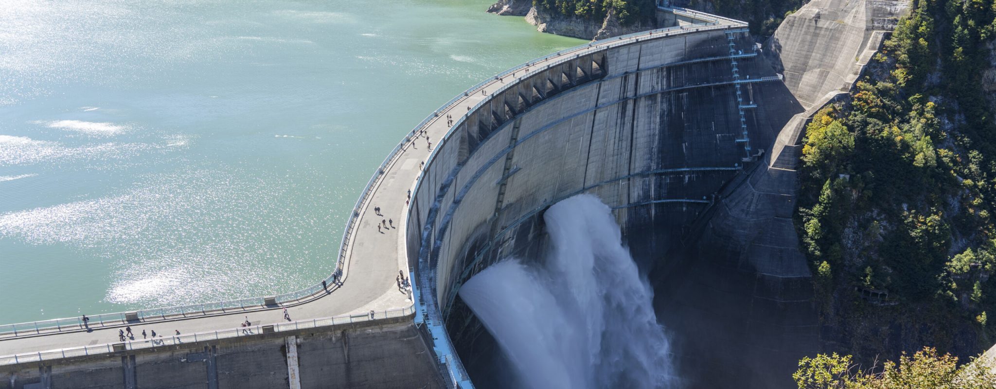 Water Turbines Are Producing Electricity At Power Plant. Panorama View Of Hydro Power Station And People On The Kurobe Lake Dam,Toyama. River Dam and Tateyama Mountains.