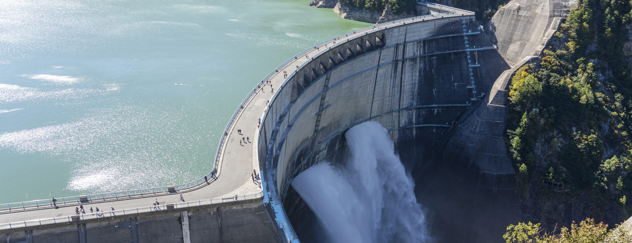 Water Turbines Are Producing Electricity At Power Plant. Panorama View Of Hydro Power Station And People On The Kurobe Lake Dam,Toyama. River Dam and Tateyama Mountains.